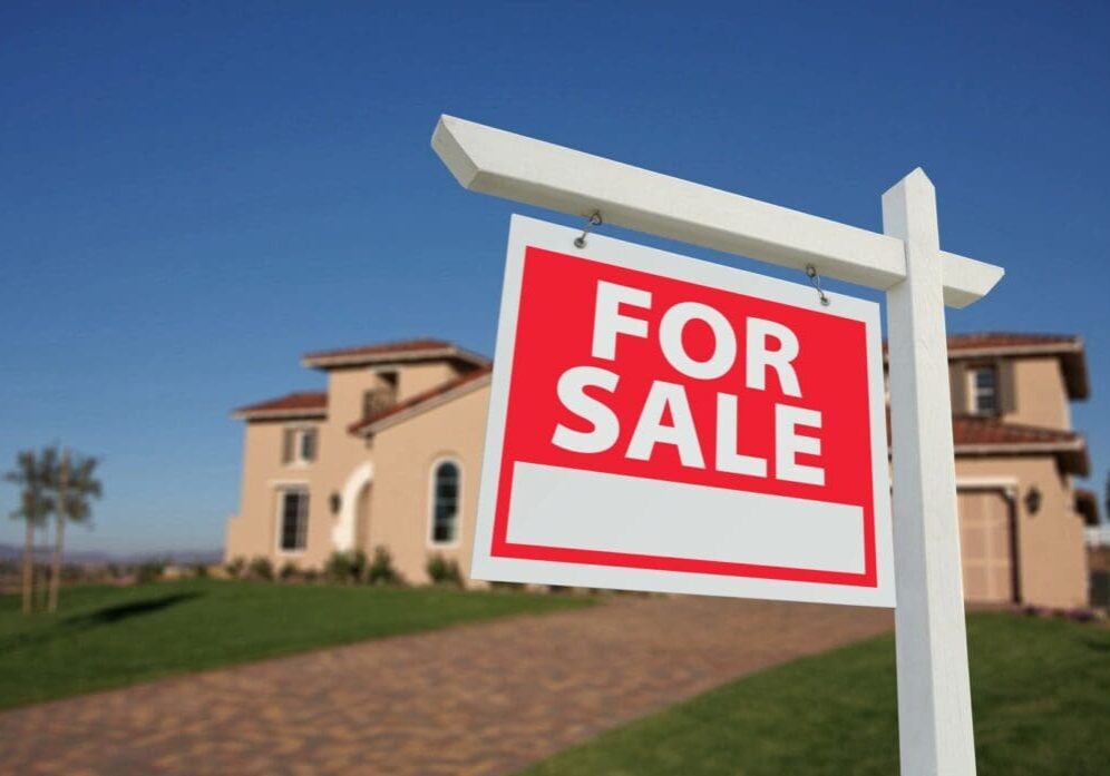 qtq80-E5kMsG "For Sale" sign in sharp focus with a blurred home in the background, representing uncertainty in the 2025 housing market.